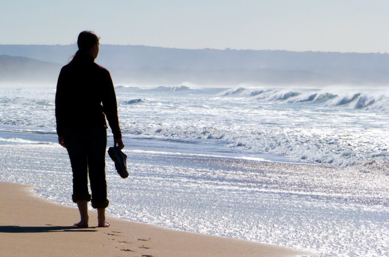 The silhouette of a woman holding her shoes and walking along the edge of the water on a sandy beach