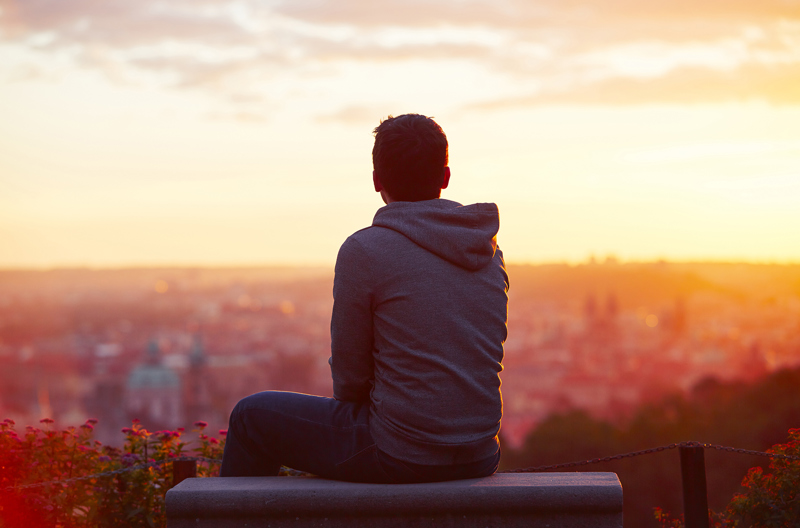 A man sitting on a bench on a hill and watching a sunset over a city
