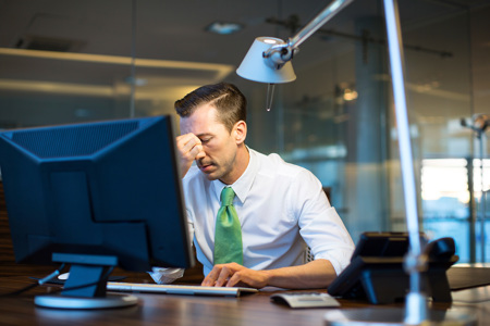 A man wearing a white shirt with a green tie sitting at a desk with a disgruntled look on his face.